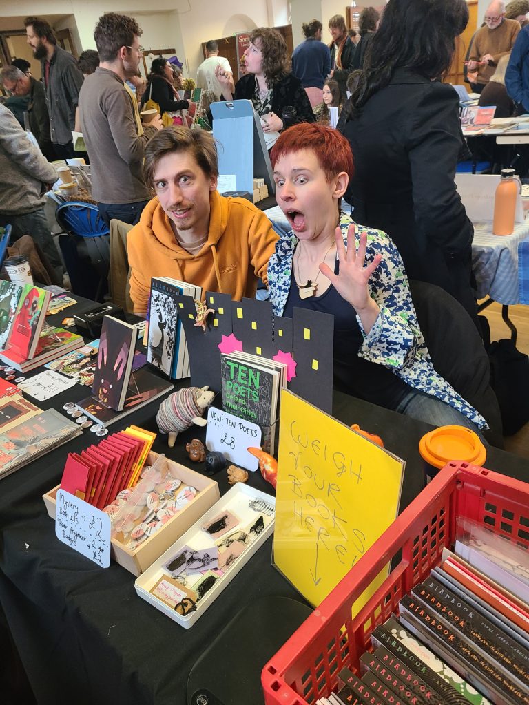 A man and a woman sitting pulling funny faces at a stall full of books.
